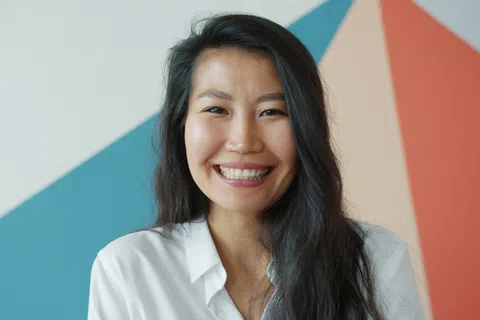 Smiling woman with long black hair wearing a white shirt against a colorful geometric wall background