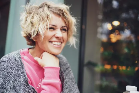 Smiling young woman with curly blonde hair wearing a pink sweater and gray cardigan sitting outdoors.