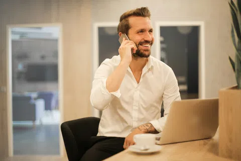 Smiling man in white shirt talking on phone while sitting at desk with laptop and coffee cup in modern office.