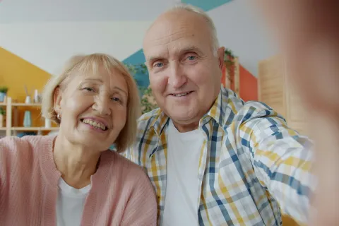 Smiling elderly couple taking a selfie together in a cozy, colorful living room setting.