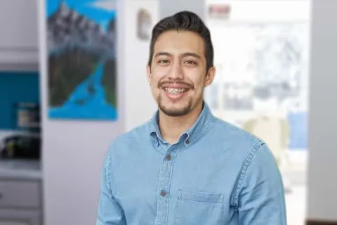 Smiling man with braces wearing a light blue denim shirt standing indoors with blurred background.