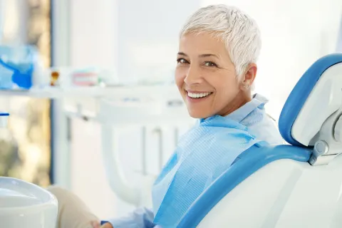Smiling older woman in a dental office, seated in a chair with dental tools visible.
