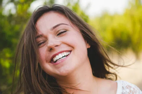 Happy young woman with brown hair smiling outdoors wearing a white lace top with greenery background