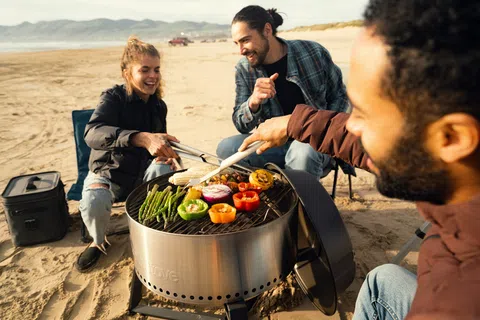 Three friends grilling mixed vegetables including peppers, corn, and asparagus on a portable grill at the beach.