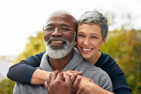 Smiling middle-aged couple outdoors, woman embraces man with grey beard and glasses on a bright day