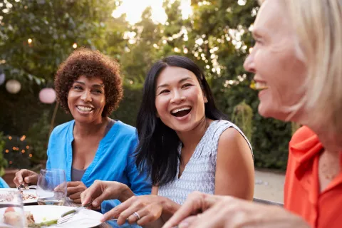 Three diverse women enjoying an outdoor meal together, laughing and sharing a joyful moment in a garden setting.