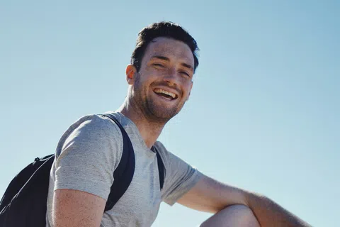 Smiling man in gray t-shirt and black shorts sitting on a rock outdoors wearing a backpack and watch on a sunny day