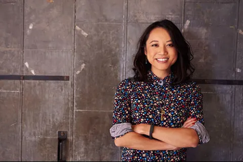 Smiling woman with folded arms wearing a colorful polka dot shirt standing against a textured metal wall.