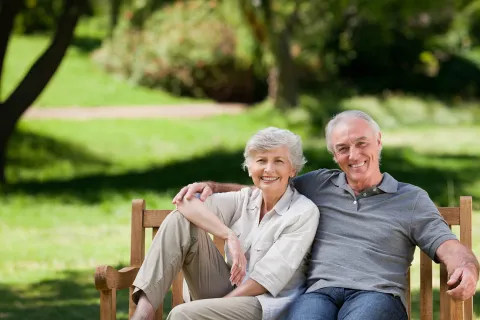 Smiling senior couple sitting on a bench in a park, enjoying a sunny day surrounded by greenery.