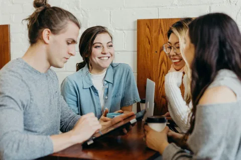 Group of young adults collaborating and smiling while using a tablet and laptop in a cafe setting.