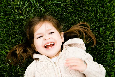 Happy young girl lying on green grass smiling and laughing with pigtails and cream sweater
