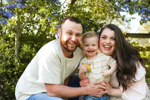 Smiling young family of three outdoors with green foliage and purple flowers in background on sunny day