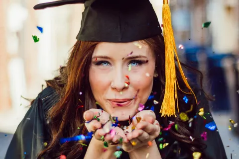 Young woman in graduation cap and gown joyfully blowing colorful confetti celebrating her achievement outdoors.