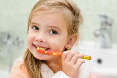 Young girl with blonde hair brushing her teeth with an orange toothbrush in a bathroom.