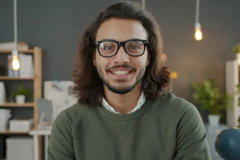 Smiling man with glasses and long hair in a cozy, modern office with shelves and hanging light bulbs.