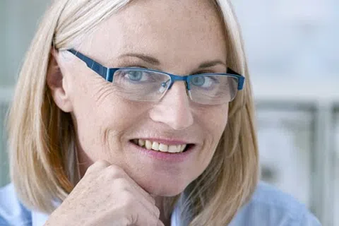 Smiling middle-aged woman with glasses and blonde hair resting her chin on her hand against a blurred background