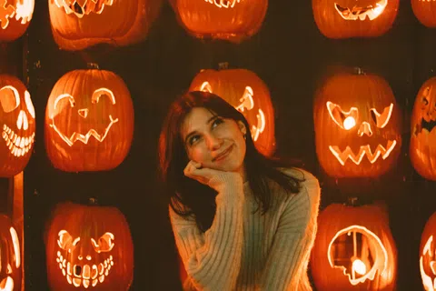 Young woman sitting thoughtfully among illuminated carved Halloween pumpkins on hay.