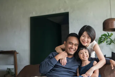 A happy family of three poses together on a couch in a cozy living room, smiling warmly at the camera.