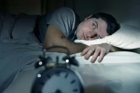 A man lying awake in bed at night, staring anxiously at an alarm clock.