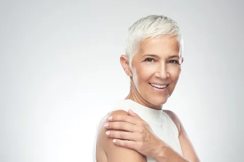 Confident elderly woman with short white hair smiling against a soft white background.