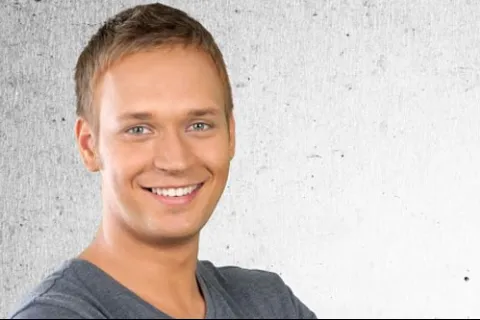 Smiling young man in gray shirt against a textured light background, conveying positivity and friendliness.