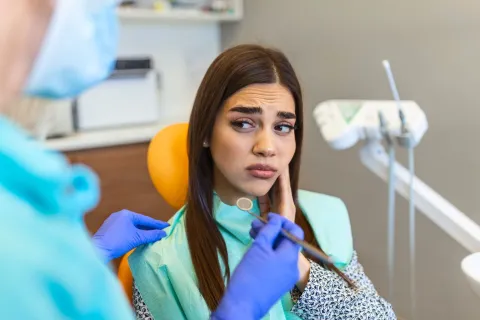 Worried woman at dentist with dentist examining her teeth and dental tools visible.