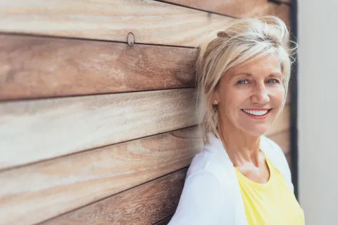Smiling mature woman with blonde hair leans against a wooden wall wearing a yellow shirt and white cardigan.