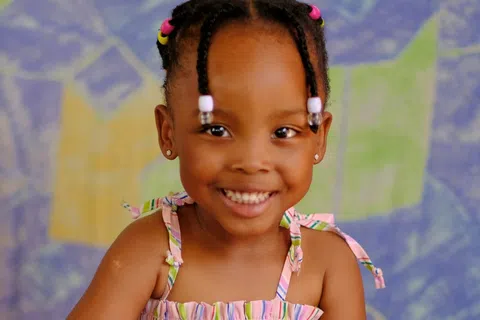 Smiling young girl in a colorful striped dress sitting with hands clasped against a vibrant abstract background.