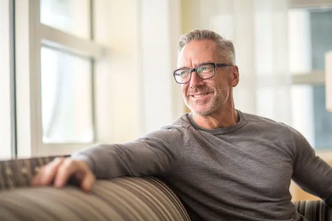 Smiling middle-aged man in glasses relaxing on a sofa by a window.