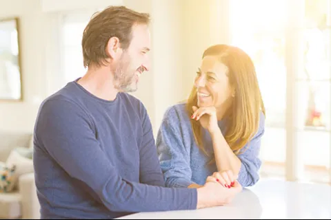 Middle-aged couple smiling and holding hands while sitting at a kitchen counter in bright sunlight
