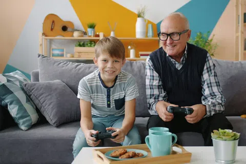 Grandfather and grandson sitting on sofa playing video games together smiling in colorful living room