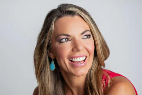 Smiling woman with long blonde hair wearing turquoise earrings and a red sleeveless top, showcasing a bright smile.