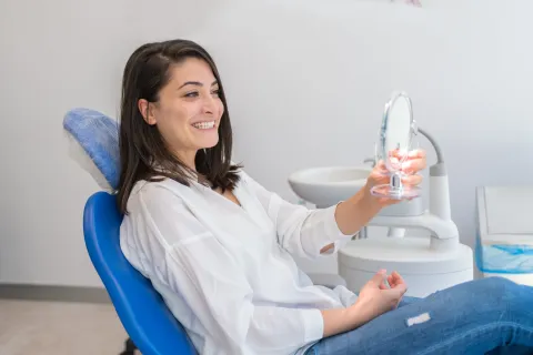 Smiling woman in dental chair examining her teeth in a mirror at a dental office.