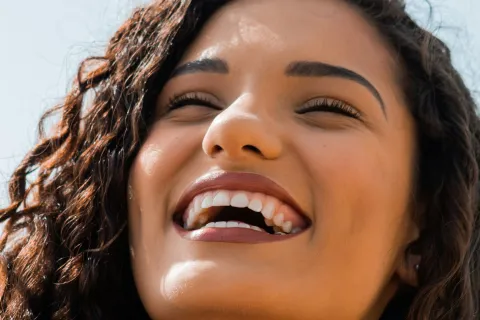 Close-up of a young woman with curly hair smiling brightly against a clear blue sky background.