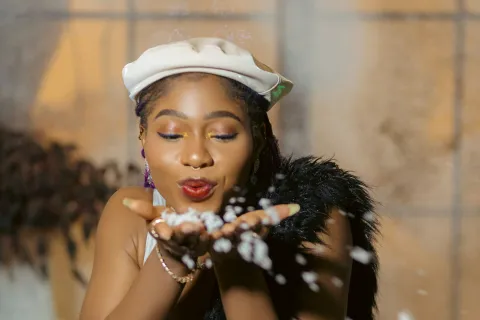 Woman in white outfit and beret blows snowflakes from hands against a softly lit window background.