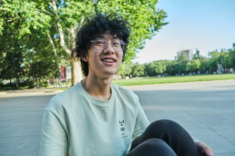 Young man with glasses and curly hair smiles outdoors in a sunny park setting wearing a light green shirt.