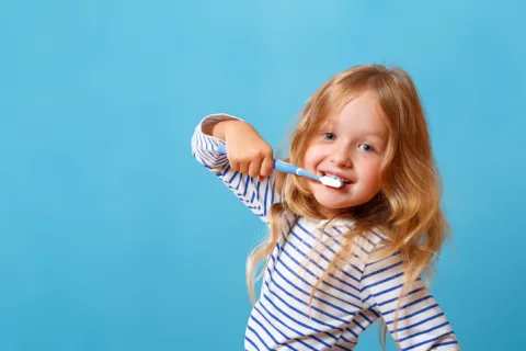 Young girl with long hair brushing her teeth against a bright blue background.