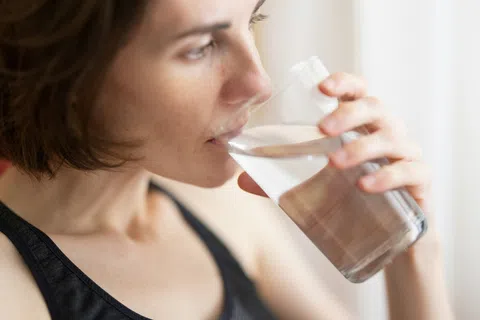 Woman in a black tank top drinking a glass of clear water indoors near a bright window.