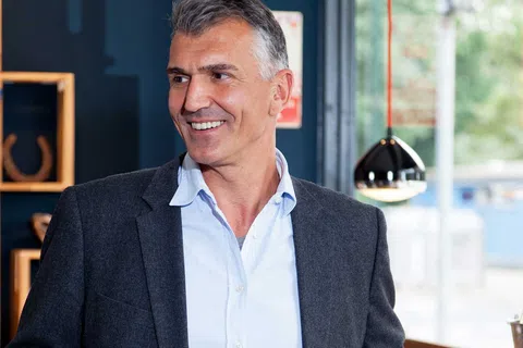 Smiling mature man in a gray blazer and blue shirt standing indoors near a window with modern decor.