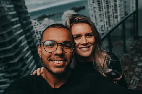 Smiling couple taking a selfie on a city balcony with wine glass and high-rise buildings in the background