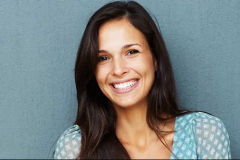 Woman with long hair smiling against a gray wall, wearing a turquoise patterned blouse.