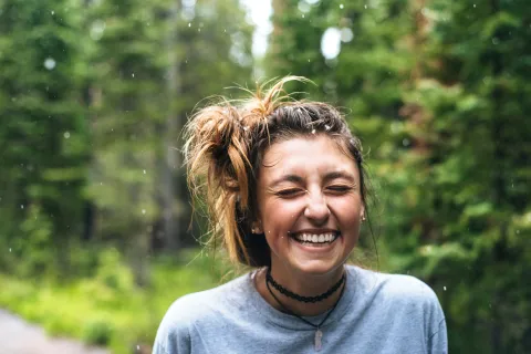 Young woman laughing with eyes closed outdoors in a forest setting with raindrops falling around her.