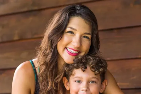 A smiling mother embraces her son, both showing joy in a cozy wooden space with colorful decor.