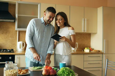 Couple preparing healthy food in kitchen using tablet for recipe while smiling and chopping vegetables