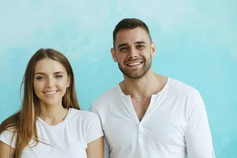 Smiling young man and woman in white shirts standing in front of a bright blue background together.