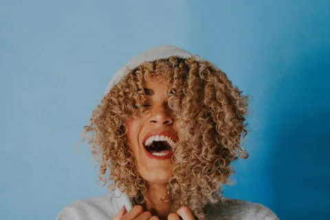 Smiling woman with curly hair wearing a grey hoodie against a blue background laughing joyfully