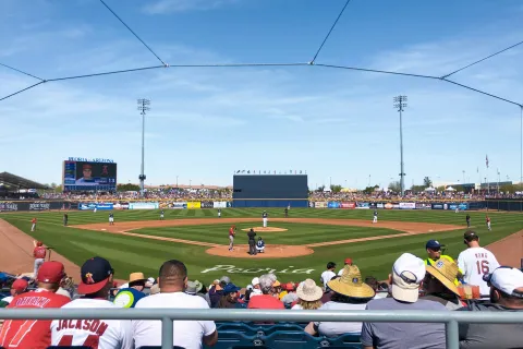 Baseball game in progress at Peoria stadium with fans watching under clear blue sky and bright sunlight