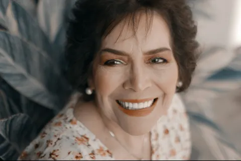 Smiling mature woman with short dark hair wearing floral blouse and pearl earrings in soft natural light.