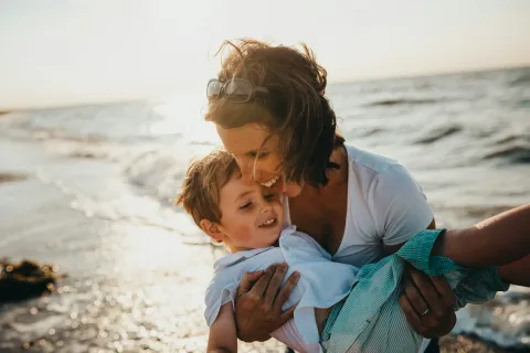 Woman holding and smiling with young boy at the beach during sunset near the ocean shore.