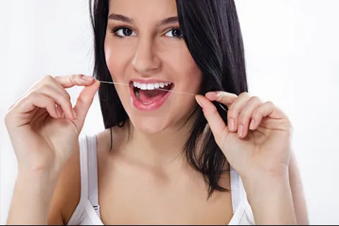 Young woman with dark hair flossing teeth, smiling with white teeth against a white background.